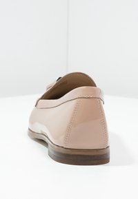 Chaussure plate en cuir verni nude avec un bout arrondi, un design simple et une semelle marron. Présente des coutures le long des bords et un matériel minimal.