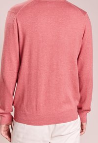 Homme portant un pull à manches longues rose et un pantalon blanc, vu de dos et debout devant un fond uni.