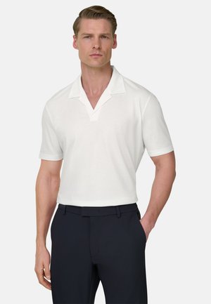 Mand med kort hår iført en hvid skjorte med korte ærmer og mørke bukser, står med den ene hånd i lommen mod en ensfarvet baggrund.