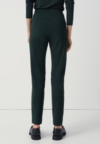 Dopasowane, ciemnozielone spodnie o gładkiej fakturze, wysokim stanie i dwóch tylnej kieszeniach. Noszone z czarnymi butami na grubych podeszwach.