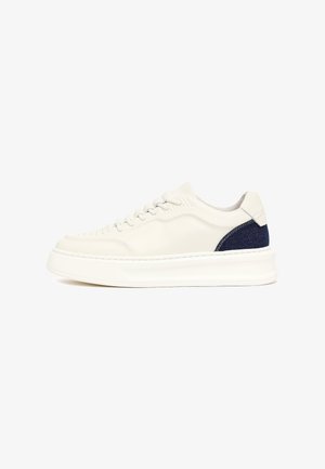 Zapatilla blanca de perfil bajo con suela gruesa y detalle en tela azul marino en el talón, vista de lado sobre fondo blanco.
