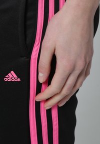 Main reposant sur un pantalon Adidas noir avec des rayures et un logo roses, mettant en valeur les détails de la tenue de sport et la texture du tissu.