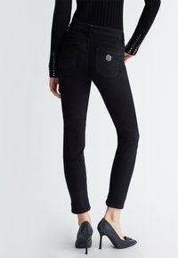 Jeans skinny in denim nero con vita alta, dotati di una tasca posteriore con logo di strass, abbinati a scarpe con tacco alto nere.