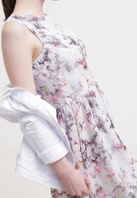 Kobieta w bezrękawnej, kwiatowej sukience z wcięciem w talii, częściowo zakładająca lub zdejmująca białą długą kurtkę.