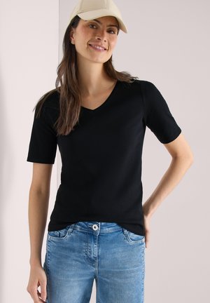 Femme portant une casquette beige, un t-shirt noir à manches courtes et col en V, et un jean bleu, souriant avec une main sur la hanche.