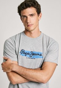 Siva pamučna majica sa plavim i crnim logotipom koji kaže "Pepe Jeans London." Kratki rukavi i okrugli izrez, glatka tekstura.
