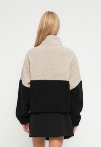 Γυναίκα με μακριά ξανθά μαλλιά που φοράει μια μπεζ και μαύρη φλις ζακέτα και μαύρη φούστα, όρθια με την πλάτη προς το θεατή, σε ένα απλό φόντο.