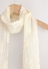 Vezen šal v svetlo kremni barvi z cvetličnimi vzorci in robom iz čipke s škrlatastim robom. Izdelan iz lahkega, prosojnega blaga.