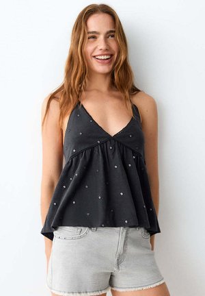Jeune femme souriante portant un top noir sans manches à petits pois et un short en jean gris clair sur fond blanc.