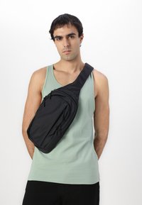 Crni crossbody ruksak s više zatvorenih džepova, napravljen od izdržljive tkanine. Nosi se preko ramena, ima glatke linije i moderan dizajn.