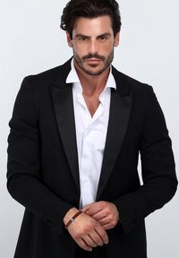 Giacca da smoking nera con rever in raso sopra una camicia bianca. Braccialetto intrecciato marrone al polso. Sfondo semplice e pulito.