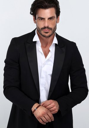 Schwarze Smokingjacke mit Satinrevers über einem weißen Hemd. Brauner geflochtener Armreif am Handgelenk. Einfacher, klarer Hintergrund.