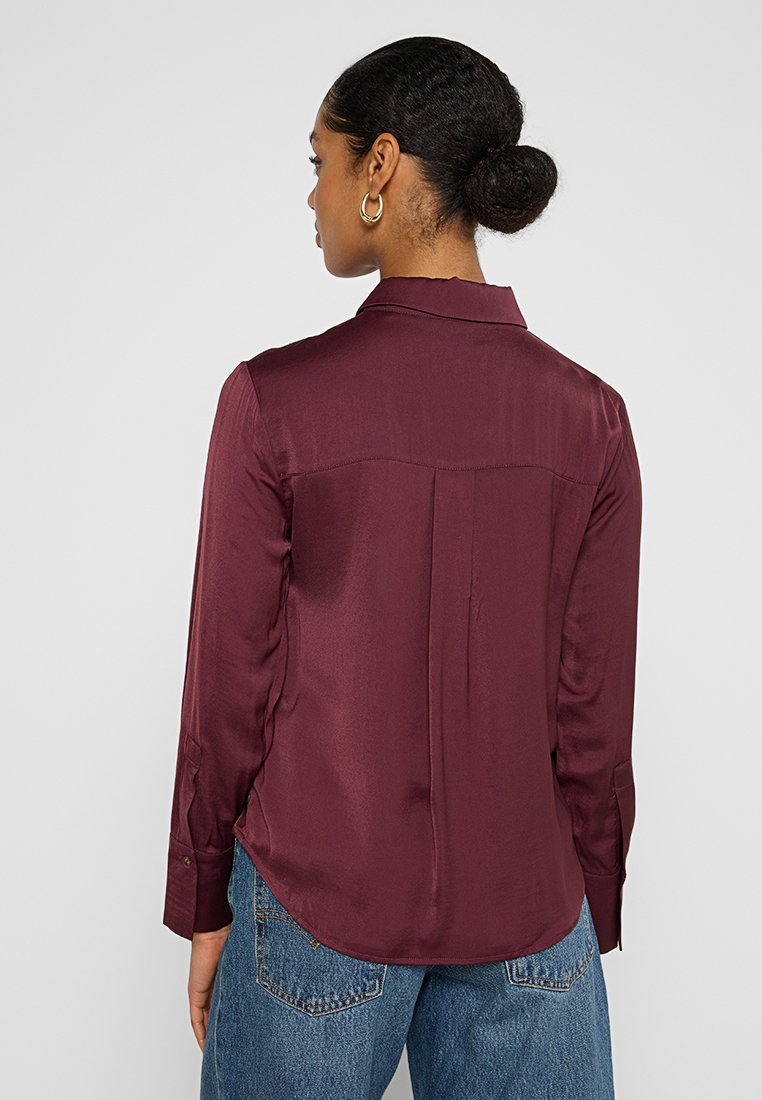 Bordeauxfarbenes Longsleeve-Hemd mit Kragen, glatter Textur und Rückenfalten-Detail, kombiniert mit lockeren Jeans.