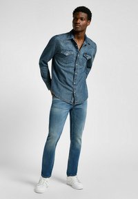 Granatowa koszula dżinsowa z zatrzaskami, spiczastym kołnierzykiem i dwoma kieszeniami na piersi; zestawiona z jasnoniebieskimi dżinsami i białymi sneakersami.