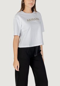 Witte gecroppte t-shirt met een gouden "ARMANI" logo, korte mouwen en een ontspannen pasvorm, gecombineerd met zwarte jogger-achtige broeken.