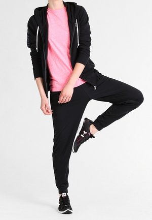 Osoba ubrana w różową koszulę, czarną bluzę z kapturem na zamek, czarne spodnie dresowe i czarne tenisówki stojąca na jednej nodze.