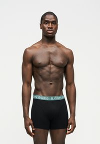 Μυώδης άνδρας στέκεται μπροστά με φορά προς τα εμπρός, φορώντας μαύρα εσώρουχα boxer Björn Borg με πράσινη ζώνη μέσης, μπροστά σε λευκό φόντο.