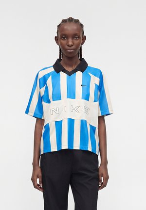 Osoba s pletenicama koja nosi plavo-bijelu Nike majicu s vertikalnim prugama, crnim ovratnikom i crne hlače, stoji ispred jednobojne pozadine.