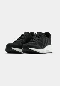 Paire de chaussures de sport noires avec une tige en mesh texturé, lacets noirs et semelles épaisses blanches sur un fond gris clair uni.