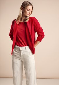 Červený kardigan přes červený top, spárovaný s lehkými béžovými širokými kalhotami. Kardigan má jemnou texturu a design s otevřeným předním dílem a ohrnutými rukávy.