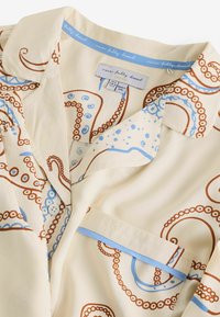 Cămașă de culoare crem, cu un model mare de caracatiță în nuanțe de maro și albastru. Gulerul are un accent de albastru deschis și include un buzunar la piept.