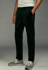 Pantaloni din catifea neagră cu croială dreaptă, având buzunare laterale și închidere cu nasture, asortați cu adidași gri.