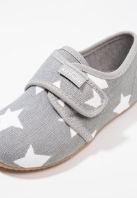 Chaussure en tissu gris avec un motif d'étoiles blanches, fermeture par sangle ajustable, surface texturée et semelle en caoutchouc marron. Design léger et décontracté.