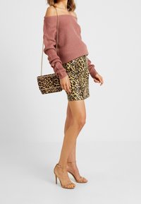 Femme portant un pull rose à épaules dénudées, une jupe à imprimé léopard, un sac à main assorti à imprimé léopard, et des sandales à talons hauts nude, debout de profil.