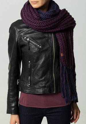 Écharpe en grosse maille violet et bleu marine drapée sur une veste en cuir noire avec poches zippées et coupe ajustée. Texture douce, aspect chaud.