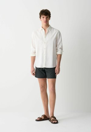 Camicia bianca in lino con bottoni, pantaloni da bagno neri e sandali marroni con fasce. Il modello si trova su uno sfondo semplice. Outfit estivo casual.