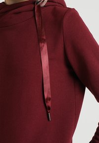 Sudadera con capucha burdeos de textura suave, con cordón de cinta de satén y terminal metálico, que destaca un diseño moderno.