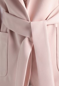 Ljusrosa bälte omfamning med slät yta, breda slag och framfickor, som betonar en ren, minimalistisk design.