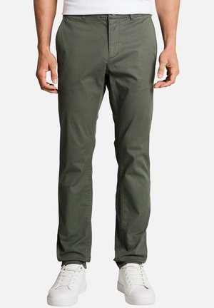 Homme portant un pantalon droit vert olive et des baskets blanches, debout devant un fond clair uni.