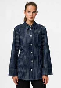 Camisa de mezclilla oscura de botones con rayas blancas verticales, que presenta un collar, dos bolsillos en el pecho y mangas anchas. Acabado de tejido texturizado.