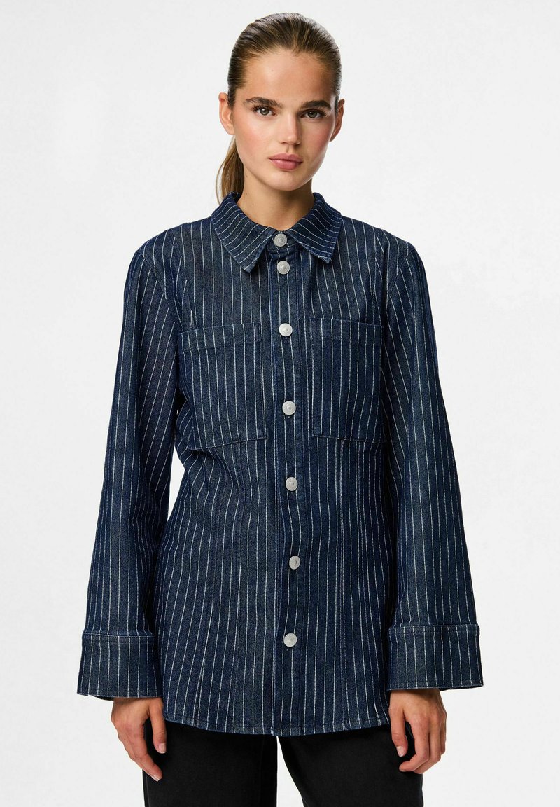 Camisa de mezclilla oscura de botones con rayas blancas verticales, que presenta un collar, dos bolsillos en el pecho y mangas anchas. Acabado de tejido texturizado.