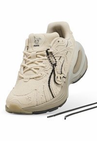 Baskets beige Puma en tissu texturé avec des détails noirs, une étiquette personnage Mimikyu et des lacets noirs détachés sur fond blanc.