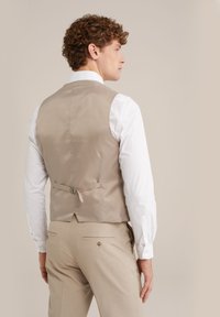 Beige Satinweste mit taillierter Passform und verstellbarem Rückengurt, kombiniert mit einem weißen Langarmhemd und hellbeigen Hosen.
