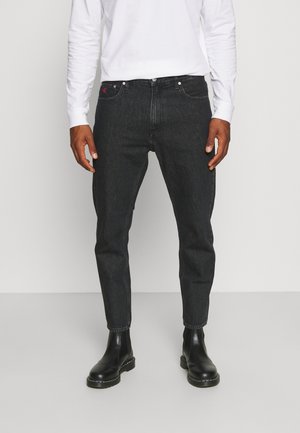 Un homme porte un jean noir ajusté et des bottes en cuir noires, associés à une chemise blanche à manches longues, debout devant un fond clair uni.