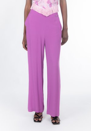 Kvinde iført lilla bukser med høj talje og vide ben med sidelommer og sorte sandaler med stropper, stående mod en hvid baggrund.