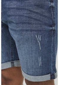 Shorts in denim con lavaggio blu scuro, dotati di una texture leggermente vissuta e orlo arrotolato. Progettati con una vestibilità attillata e dettagli di cucitura in evidenza.