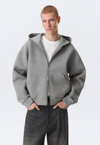 Junger Mann mit kurzen Haaren, der einen grauen Kapuzenpullover mit Reißverschluss über einem weißen Hemd und dunkelgrauen Hosen trägt und mit den Händen in den Taschen posiert.