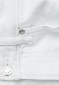Close-up van witte denimstof met een riemlus, zichtbare stiksels en twee zilveren metalen klinknagels met gegraveerde tekst.