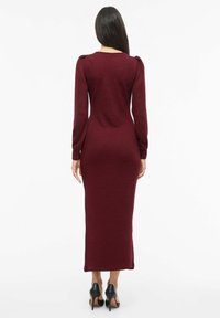 Mujer con cabello largo y oscuro, vestida con un vestido midi ajustado de manga larga color burdeos y tacones altos negros, mirando de espaldas sobre un fondo blanco.