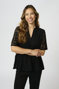 Blusa nera in pizzo con maniche corte, scollatura a V e design a peplum. Presenta motivi floreali e una texture morbida. Indossata con pantaloni neri.