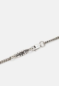 Pulseira de corrente de prata com uma etiqueta oval texturizada que lê "CLASSICS 77" e um fecho de lagosta. Apresenta elos e um acabamento metálico liso.