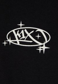 Sort stof med et hvidt logo med teksten "k1x", omkranset af dekorative stjerner og en oval kontur. Designet er dristigt og grafisk.