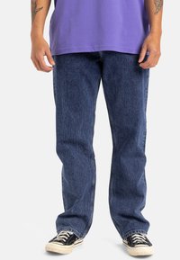 Vaqueros de denim azul con un corte relajado, que presentan una textura sutil y un diseño clásico de cinco bolsillos, combinados con zapatillas en blanco y negro.