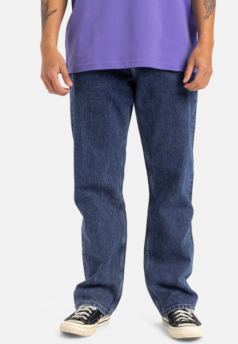 Vaqueros de denim azul con un corte relajado, que presentan una textura sutil y un diseño clásico de cinco bolsillos, combinados con zapatillas en blanco y negro.