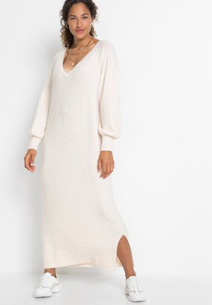 Robe longue en tricot blanc cassé avec un décolleté en V, des manches ballon et une fente sur le côté, associée à des baskets blanches. Texture douce, coupe décontractée.
