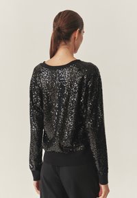 Schwarzer Paillettenpullover mit Rundhalsausschnitt, langen Ärmeln und gerippten Bündchen. Die Pailletten bedecken den Stoff und erzeugen ein strukturiertes, glänzendes Erscheinungsbild.